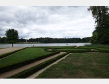 Ein gepflegter Park mit symmetrischen Hecken und Wegen grenzt an einen ruhigen See. Im Hintergrund sind dichte Wälder zu sehen, und auf dem Wasser befinden sich Boote; links steht ein einzelner Baum.