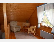 Wohnraum in einem Dachgeschoss mit Holzvertäfelung. Die Einrichtung besteht aus einem gepolsterten Sessel, zwei Korbstühlen, einem kleinen runden Tisch und einem Fenster mit weißen Vorhängen, die blau eingefasst sind.