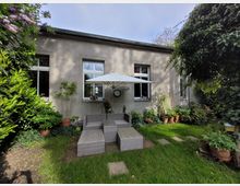 Kleiner Gartenbereich mit einer hellgrauen Hausfassade und drei großen Fenstern. Im Vordergrund stehen Gartenmöbel aus Polyrattan und ein weißer Sonnenschirm, umgeben von verschiedenen Topfpflanzen und dichtem Grün.