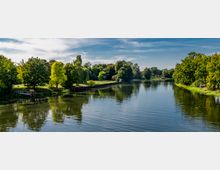 Flusslandschaft mit ruhigem Wasser, umgeben von dichtem, grünem Baumbestand und Rasenflächen; im Hintergrund blauer Himmel mit vereinzelten Wolken. Entlang des Ufers sind befestigte Böschungen und vereinzelte Parkelemente erkennbar.