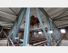 Innenansicht eines historischen Gebäudes mit sichtbaren Stahlkonstruktionen und Holz-Elementen. Ziegelwände, große Fenster und ein hölzernes Dachgebälk prägen den Raum, während Galerien und Metallstreben die Struktur stützen.