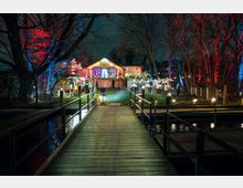 Holzsteg führt zu einem beleuchteten Gebäude mit großen Fenstern, umgeben von Gartenbereichen und Terrassen mit dekorativen Lichtern. Im Vordergrund befinden sich Bäume, teilweise in roten und blauen Farben angestrahlt, die eine stimmungsvolle Atmosphäre schaffen.