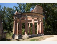 Ruine eines Rundtempels aus rotem Backstein, bestehend aus mehreren Säulen und teilweise erhaltenen Wänden, umgeben von Bäumen in einer parkähnlichen Landschaft. Der Boden ist grasbedeckt, teils mit einem Weg daneben.