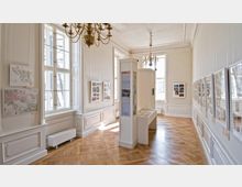 Heller Ausstellungsraum mit Holzfußboden, großen Fenstern und weißen Wänden, an denen Karten und Bilder in Rahmen hängen. In der Raummitte stehen hohe Vitrinen mit Text- und Bildinformationen.