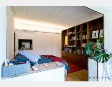 Modernes Schlafzimmer mit integriertem Wohnbereich, einer großen Holzregalwand voller Bücher und Dekorationen auf der rechten Seite. Im Vordergrund ein Bett mit blauer und roter Bettwäsche sowie ein kleiner Tisch mit diversen Gegenständen; links schlichte weiße Wände und Schranktüren, eine Pflanze steht rechts vorne.