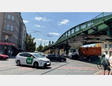Straßenszene an einer Kreuzung in einer Stadt mit vorbeifahrenden Autos, einem Fahrradfahrer und Fußgängern. Im Hintergrund sind ein graues Wohngebäude, eine grüne Hochbahnbrücke und der Fernsehturm sichtbar.