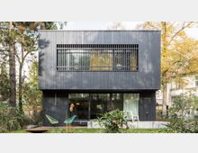 Modernes Wohnhaus mit dunkler Holzfassade, großem Fensterelement im Erdgeschoss und vertikalen Lamellen vor einem Fenster im Obergeschoss. Das Gebäude steht in einem Garten mit Pflanzen und Bäumen, umgeben von weiteren Wohnhäusern.