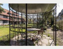 Ein Gartenpavillon mit Metallstruktur, ausgestattet mit einem Tisch und mehreren Holzstühlen, steht in einem großzügigen, grünen Garten. Im Hintergrund ist ein großes Haus mit rotem Ziegeldach und mehreren Fenstern zu sehen, umgeben von Bäumen und Beeten.