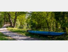 Ein schmaler Weg führt durch einen von Bäumen gesäumten Park neben einem ruhigen Fluss. Im Vordergrund liegt ein blaues Boot am Ufer, während im Hintergrund eine kleine Brücke mit Geländer über den Fluss führt.