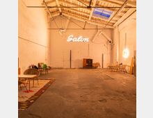 Ein industriell gestalteter Raum mit hoher Decke und sichtbaren Holzbalken, weiß gestrichenen Backsteinwänden und einem Betonboden. Möbel, darunter Stühle und Tische, sind vereinzelt aufgestellt, und an der Rückwand hängt ein leuchtendes Neon-Logo mit der Aufschrift „Salon“.