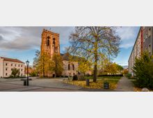Historische Backsteinkirche mit hohem Turm in einer herbstlichen Umgebung, umgeben von Laubbäumen, einem gepflasterten Platz und Wohngebäuden.