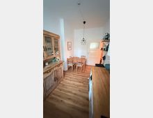 Schmale Küche mit Holzfußboden und rustikalen Holzmöbeln. Links steht eine Vitrine, rechts eine Arbeitsplatte mit Regalen darüber, im hinteren Bereich ein Esstisch mit vier Stühlen und eine Tür.