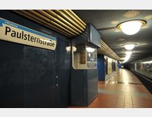 U-Bahn-Station "Paulsternstraße" mit blauen Wänden, einer Lichtdecke mit runden Lampen und goldenen Verzierungen sowie einem gewölbten Bahnsteig aus roten Fliesen. Im Hintergrund sind Gleise und Wandmalereien zu sehen.