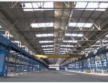 Große, leere Industriehalle mit einer Konstruktion aus Stahlträgern und Oberlichtern, die Tageslicht hereinlassen. Die Halle verfügt über blaue Stahlstützen und eine gelbe Laufkrantraverse in der Mitte.