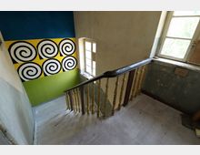 Treppenhaus in einem alten Gebäude mit abgenutztem Holzgeländer und hellen Holzdielenstufen. An der Wand ein farbiges Wandbild mit spiralförmigen Mustern vor gelbem und grünem Hintergrund, daneben ein Fenster mit verwittertem Rahmen.