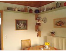 Ein gemütlicher Essbereich in einem klassischen Wohnraum, mit hellgrünen Wänden und Holzmöbeln. An der Wand hängen Regale mit dekorativen Keramik- und Glasgefäßen, eine Lampe mit drei Schirmen hängt über einem Tisch mit karierter Tischdecke, darauf ein Dekorationskrug mit gelben Blumen. Wandbilder und ein Heizkörper ergänzen die Einrichtung.