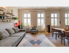 Helles Wohnzimmer mit großen, bodentiefen Fenstern, die viel Tageslicht hereinlassen. Der Raum ist mit einem grauen Sofa, einem Esstisch mit Holzstühlen, einem geometrischen Teppich und mehreren Pflanzen dekoriert; an einer Wand befinden sich offene Regale.