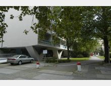 Wohngebäude mit moderner Architektur, das auf spitz zulaufenden Betonstützen steht, umgeben von Gehwegen, Bäumen und einer Grünfläche. Im Vordergrund ist ein geparkter silberner Pkw sowie eine Absperrkette mit roten Pollern zu sehen.