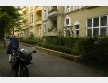 Mehrfamilienhaus im Altbaustil mit gelben Fassaden und dekorativen Elementen, umgeben von kleinen Vorgärten und Hecken. Im Vordergrund steht ein Motorrad auf einem gepflasterten Gehweg.