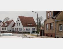 Straßenszene in einem Wohngebiet mit Einfamilienhäusern im Stil der 20er oder 30er Jahre, gekennzeichnet durch rote Satteldächer und weiß verputzte Fassaden. Im Vordergrund steht ein Verkehrsschild, ein Gehweg verläuft entlang der Straße, und vereinzelte Bäume sind im Hintergrund sichtbar.