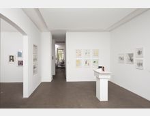 Minimalistisch gestalteter Galerieraum mit weißen Wänden und modernen Kunstwerken in schlichten Rahmen. Im Vordergrund steht ein weißer Sockel, im Hintergrund führt ein schmaler Durchgang zu einem weiteren Raum mit einem Fenster.
