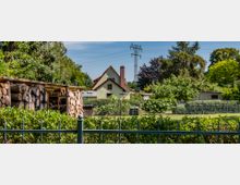 Ein Einfamilienhaus mit rotem Satteldach steht inmitten eines Gartens mit Gemüsebeeten, Sträuchern und Bäumen. Im Vordergrund befindet sich ein Stapel Holz unter einem Unterstand sowie eine Hecke mit einem Metallzaun.