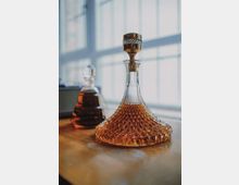 Zwei gläserne Karaffen stehen auf einem Holztisch, gefüllt mit bernsteinfarbener Flüssigkeit, im Hintergrund sind große, vom Tageslicht beleuchtete Fenster erkennbar. Die vordere Karaffe hat ein geschliffenes Muster und einen goldfarbenen Deckel.