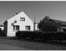Ein Einfamilienhaus mit weiß verputzter Fassade, Spitzdach und zwei Sat-Schüsseln an der Außenwand, daneben ein Ziegelgebäude. Im Vordergrund befinden sich eine Hecke, ein Maschendrahtzaun und eine Rasenfläche.