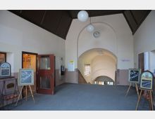 Eingangsbereich mit heller, gewölbter Decke und einer großen Uhr über einem Treppenabgang. Links befindet sich eine geöffnete Tür mit Hinweistafeln, die ein Café ankündigen.