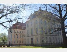 Historisches Schlossgebäude mit hellgelber Fassade und rotem Mansarddach, umgeben von kahlen Bäumen im Vordergrund. Der sonnige Park im Umfeld betont die ruhige Umgebung des Gebäudes.