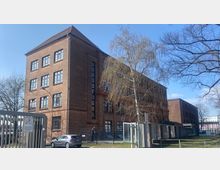 Ziegelstein-Gebäude im industriellen Stil mit mehreren Stockwerken und großen Fenstern, umgeben von einem Metallzaun. Im Vordergrund stehen ein Baum mit kahlen Ästen und geparkte Autos auf einem Gelände bei klarem Himmel.