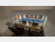 Ein Dolmetscherraum mit zwei Arbeitsplätzen, ausgestattet mit Mikrofonen, Kopfhörern und Steuergeräten, deren Fenster auf einen Veranstaltungsraum mit blauen Sitzen und einer Bühne zeigt. Die Wände des Raums sind weiß und vertikal gerillt.