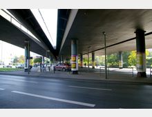 Unterführung mit breiten Autobahnbrücken, gestützt von runden Betonpfeilern, darunter Gehwege, Grünflächen und parkende Autos. Laternen und Werbung an den Pfeilern, im Hintergrund Häuser und Bäume.