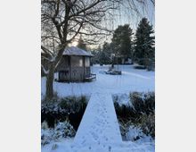 Winterliche Gartenszene mit Schnee bedecktem Boden, einem kleinen Bach und einem schmalen Holzsteg, der zu einer hölzernen Gartenhütte führt. Im Hintergrund sind Bäume und weitere Hütten zu sehen, während die tiefstehende Sonne durch die Äste scheint.