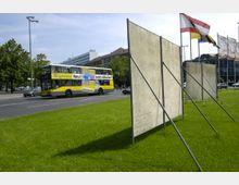 Eine städtische Straßenszene mit einer grünen Wiese im Vordergrund, auf der mehrere aufgestellte Plakatwände stehen. Im Hintergrund fahren Autos, darunter ein gelber Doppeldeckerbus mit Werbung, entlang einer Straße, während Fahnen auf Masten und moderne Gebäude im Hintergrund sichtbar sind.