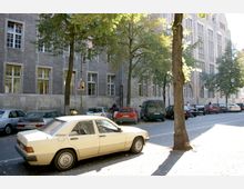 Straßenszene vor einem großen, steinernen Gebäudekomplex mit hohen Fenstern und klassischer Architektur. Auf der Straße stehen geparkte Autos, während im Vordergrund Bäume und ein Taxi zu sehen sind.