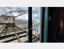 Blick durch eine Glastür auf einen historischen Industriehafen mit rostigem Hafenkran, Backsteingebäuden und Wasserwegen, bei bewölktem Himmel. Im Vordergrund ist ein Metallgerüst und ein gepflasterter Kai zu sehen.