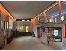 Großzügiger Innenraum eines Gebäudes mit freigelegten Ziegelwänden, sichtbaren Stahltreppen und einem Zwischengeschoss mit Glasgeländer. Der Raum ist modern beleuchtet, mit warmen und farbigen Akzentlichtern, und besitzt große Fenster, die viel Tageslicht hereinlassen.