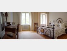 Schlafzimmer mit einem schmiedeeisernen Doppelbett und hell gemusterter Bettwäsche, zwei großen Fenstern mit weißen Vorhängen, Holzboden und einem antiken Schreibtisch mit Lampe auf der linken Seite.