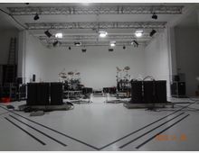Ein großer, weißer Probenraum mit mehreren Lichttraversen an der Decke, einem Schlagzeug in der Mitte und großen schwarzen Lautsprechern sowie technischen Geräten am Boden. Kabel und Musik-Equipment sind über den Raum verteilt.