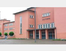 Rosa Gebäude mit der Aufschrift "Stadttheater" in schlichter, moderner Architektur. Das Gebäude hat mehrere rechteckige Fenster, Gittertore am Eingangsbereich und eine schmale Glasfensterleiste an der Frontfassade.