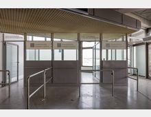 Ein Flughafen-Passkontrollbereich mit grauen Kabinen, Glasfenstern und Beschilderung "Passkontrolle/Passport control". Metallgeländer begrenzen die Warteschlangen, der Boden ist aus hellem Stein, und im Hintergrund sind Fenster mit Blick nach draußen.