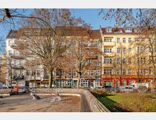 Blick auf einen kleinen Stadtplatz mit gebogener Holzsitzbank, gepflasterter Fläche und Rasenbeet mit kahlen Bäumen. Im Hintergrund stehen mehrstöckige Altbau-Wohnhäuser mit vielen Fenstern, Balkonen und farbigen Erdgeschosszonen entlang einer Straße mit geparkten Autos.