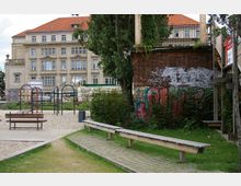 Ein kleiner Spielplatz mit Klettergerüst und Sandboden, umgeben von einer Grünfläche mit Holzsitzbänken. Im Hintergrund steht ein historisches Gebäude mit hellbeiger Fassade und rotem Ziegeldach, daneben eine Backsteinwand mit Graffiti.