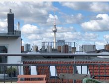 Blick über Berliner Dächer mit roten Ziegeldächern und modernen Gebäuden, im Hintergrund der Berliner Fernsehturm und das Hochhaus des Park Inn Hotels. Der Himmel ist klar mit einigen Wolken, im Vordergrund Teile einer Terrasse mit Geländer und Stühlen.