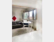 Ein minimalistischer Innenraum mit weiß gestrichenen Wänden, einem grauen Sofa, einem kleinen weißen Tisch und einem schwarzen Stuhl. Ein geöffnetes Fenster gibt den Blick auf eine städtische Skyline frei, und an der Wand hängt ein rotes, quadratisches Dekorationselement.