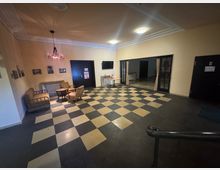 Ein Innenraum mit kariertem Boden in Schwarz und Beige, eingerichtet mit vintageartigen Sesseln und Sofas um einen kleinen Tisch. An der Wand hängen Schwarz-Weiß-Bilder, ein Kronleuchter hängt von der Decke, und eine Glastür führt in einen anderen Raum.