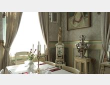 Innenraum eines elegant dekorierten Speisezimmers mit einem gedeckten Tisch, an dem ein weißes Tischtuch und Kerzenleuchter stehen. Im Hintergrund befinden sich verzierte Vorhänge, eine antike Kachelheizung, ein silberner Samowar und gerahmte Gemälde an einer mit Ornamenttapete verkleideten Wand.