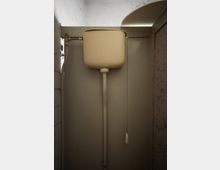 Abflusskasten in einer kleinen, engen Toilettenkabine mit beige-farbenem Wasserbehälter, einer Zugkette und sichtbaren Rohrleitungen. Die Wände sind schlicht und teilweise fleckig.