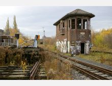 Stillgelegtes Bahngelände mit einem alten, mit Graffiti bedeckten Stellwerk aus Backstein und verwilderten Gleisen; im Vordergrund befinden sich eine Drehscheibe und überwucherte Schienen.